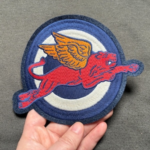 May include: Embroidered patch featuring a red winged lion in mid-flight, set against a blue and white circular background. The lion has orange wings and is detailed with intricate stitching. The patch has a dark blue border.