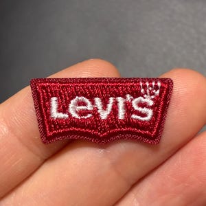 以下が含まれることがあります： スタイライズされたフォントで白い文字「Levi's」が刺繍された、小さな長方形の赤いワッペン。ワッペンは特徴的なバットウィング型で、下端が尖っており、全体に質感のあるステッチが施されています。