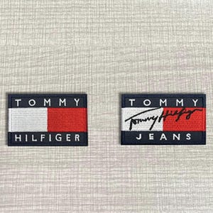 Könnte beinhalten: Zwei rechteckige gestickte Aufnäher mit dem Tommy Hilfiger Logo. Der linke Aufnäher zeigt den Markennamen in weißer Schrift auf einem rot-weißen Blockdesign. Der rechte Aufnäher zeigt das Tommy Jeans Logo mit einer schwarzen Schreibschrift.