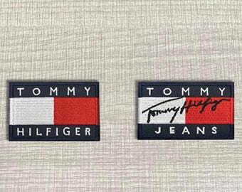 Może przedstawiać: Dwie prostokątne haftowane naszywki z logo Tommy Hilfiger. Lewa naszywka przedstawia nazwę marki białym tekstem na czerwono-białym tle. Prawa naszywka zawiera logo Tommy Jeans z czarnym podpisem wykonanym pismem odręcznym.