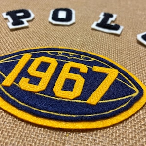 Puede incluir: Un parche de fieltro azul marino y amarillo con forma de balón de fútbol americano con el número 1967 en costura amarilla. Sobre el parche, las letras P, O, L, O están dispuestas en parches individuales de fieltro blanco y negro.