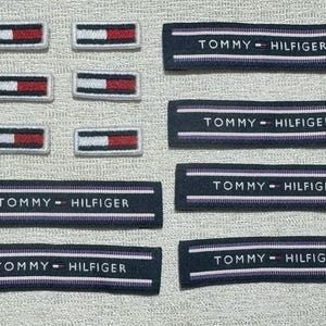 Puede incluir: Una colección de parches rectangulares de tela que incluye seis pequeños logotipos de bandera de Tommy Hilfiger en rojo, blanco y azul, junto con seis etiquetas tejidas azul marino más largas con el texto Tommy Hilfiger y finas rayas horizontales blancas y moradas.