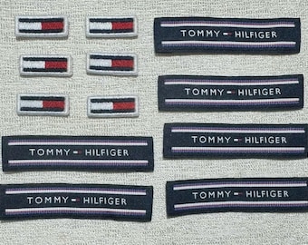 May include: A collection of rectangular fabric patches featuring six small Tommy Hilfiger flag logos in red, white, and blue, alongside six longer navy blue woven labels with the text Tommy Hilfiger and thin white and purple horizontal stripes.