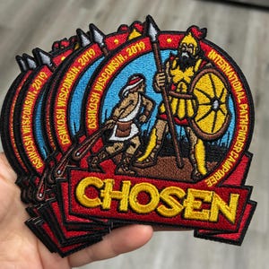 May include: A circular embroidered patch featuring a warrior in yellow armor holding a spear and shield, with a smaller figure nearby. The text reads OSHKOSH WISCONSIN, 2019, INTERNATIONAL PATHFINDER CAMPOREE, and CHOSEN. The design uses red, yellow, and blue thread.