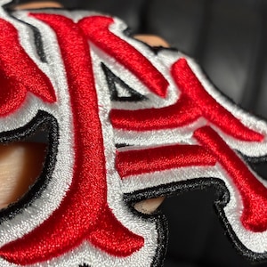 May include: A fabric patch featuring raised embroidery in bright red, white, and black thread. The design consists of stylized, interconnected lettering with a thick red center, a white border, and a thin black outline, creating a layered effect.