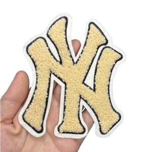 May include: A gold-colored, embroidered patch in the shape of the letters NY, with a black outline and a white border. The patch has a textured, fuzzy appearance.