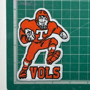 May include: Embroidered patch featuring a cartoon football player in an orange uniform, holding a football. The player is running, with the word "VOLS" in orange letters below. The patch has a white border and is on a green surface.