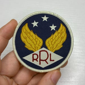 Puede incluir: Un parche redondo con fondo azul marino, con dos alas doradas, tres estrellas blancas y las letras "RRL" en rojo dentro de un rombo blanco. El parche tiene un borde blanco.