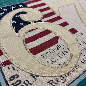 Puede incluir: Un parche de tela con un gran número seis bordado en beige sobre un fondo de la bandera estadounidense y el texto impreso 101st Cavalry U.S. Div. El diseño incluye rayas rojas y blancas y un campo azul con estrellas blancas.