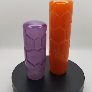 May include: Two cylindrical gear shift knobs with a textured honeycomb pattern. One knob is a translucent purple, and the other is a solid orange. Both items feature a rounded top and a smooth base, standing upright on a circular black platform.