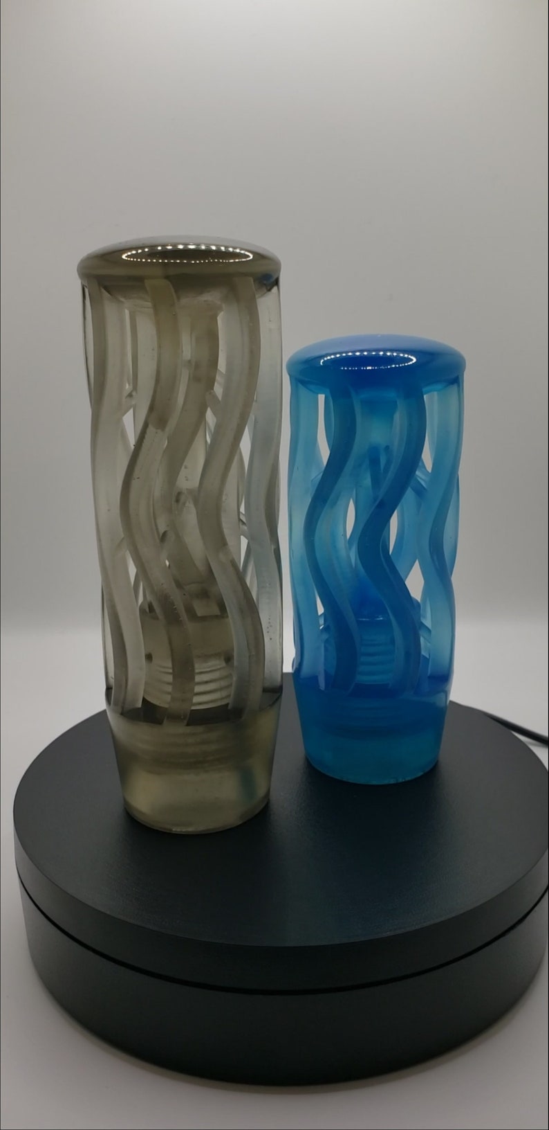 May include: Two cylindrical objects with a translucent finish, one in a smoky gray and the other in a vibrant blue. Each piece features a series of vertical, wavy, open-work structural elements that create a cage-like appearance around a central core.
