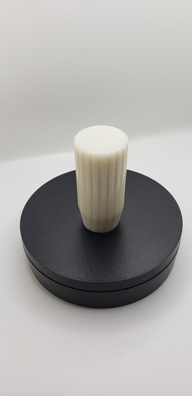 May include: A cylindrical off-white knob featuring a vertical ribbed texture along its sides and a smooth, flat circular top. The object is positioned upright, showcasing its uniform color and consistent fluted pattern throughout the exterior surface.