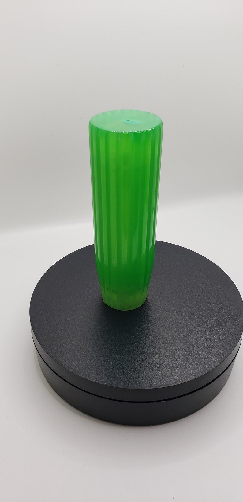 May include: A vibrant, translucent green cylindrical handle featuring a vertical ribbed texture along its length. The object has a flat, circular top surface and a slightly tapered base, showcasing a smooth, glossy finish throughout its structure.