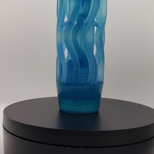 May include: A translucent blue cylindrical object featuring a series of vertical, wavy, ribbon-like structures that spiral around a central core. The piece has a smooth, rounded top cap and a solid, slightly tapered base, creating a cage-like appearance.