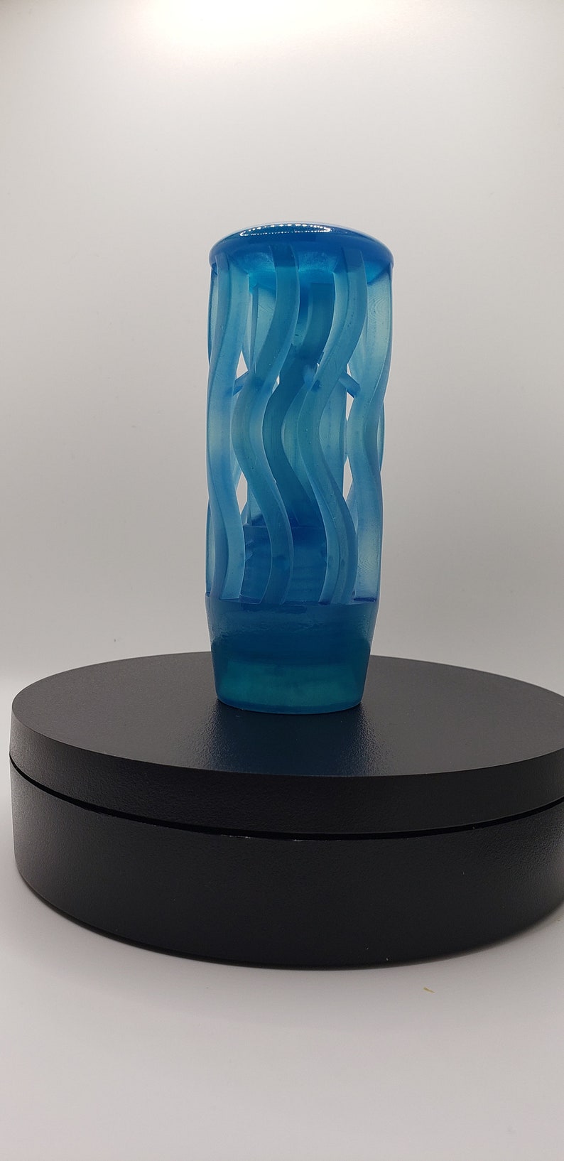 May include: A translucent blue cylindrical object featuring a series of vertical, wavy, ribbon-like structures that spiral around a central core. The piece has a smooth, rounded top cap and a solid, slightly tapered base, creating a cage-like appearance.