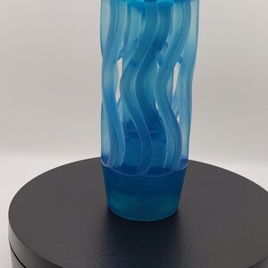 May include: A translucent blue gear shift knob featuring a cylindrical shape with a series of vertical, wavy, cut-out channels that spiral slightly around the body. The top is smooth and rounded, while the base is solid and tapers inward slightly.