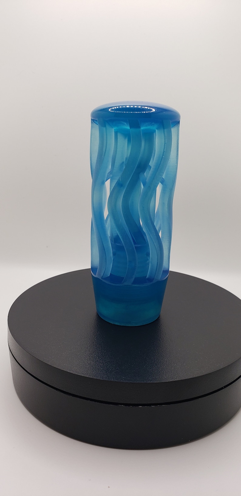 May include: A translucent blue gear shift knob featuring a cylindrical shape with a series of vertical, wavy, cut-out channels that spiral slightly around the body. The top is smooth and rounded, while the base is solid and tapers inward slightly.