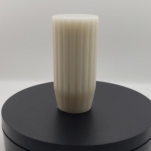 May include: A cylindrical off-white vase featuring a vertical ribbed texture along its exterior. The vessel has a flat top opening and a slightly tapered base, presenting a clean, uniform appearance with a consistent matte finish throughout.