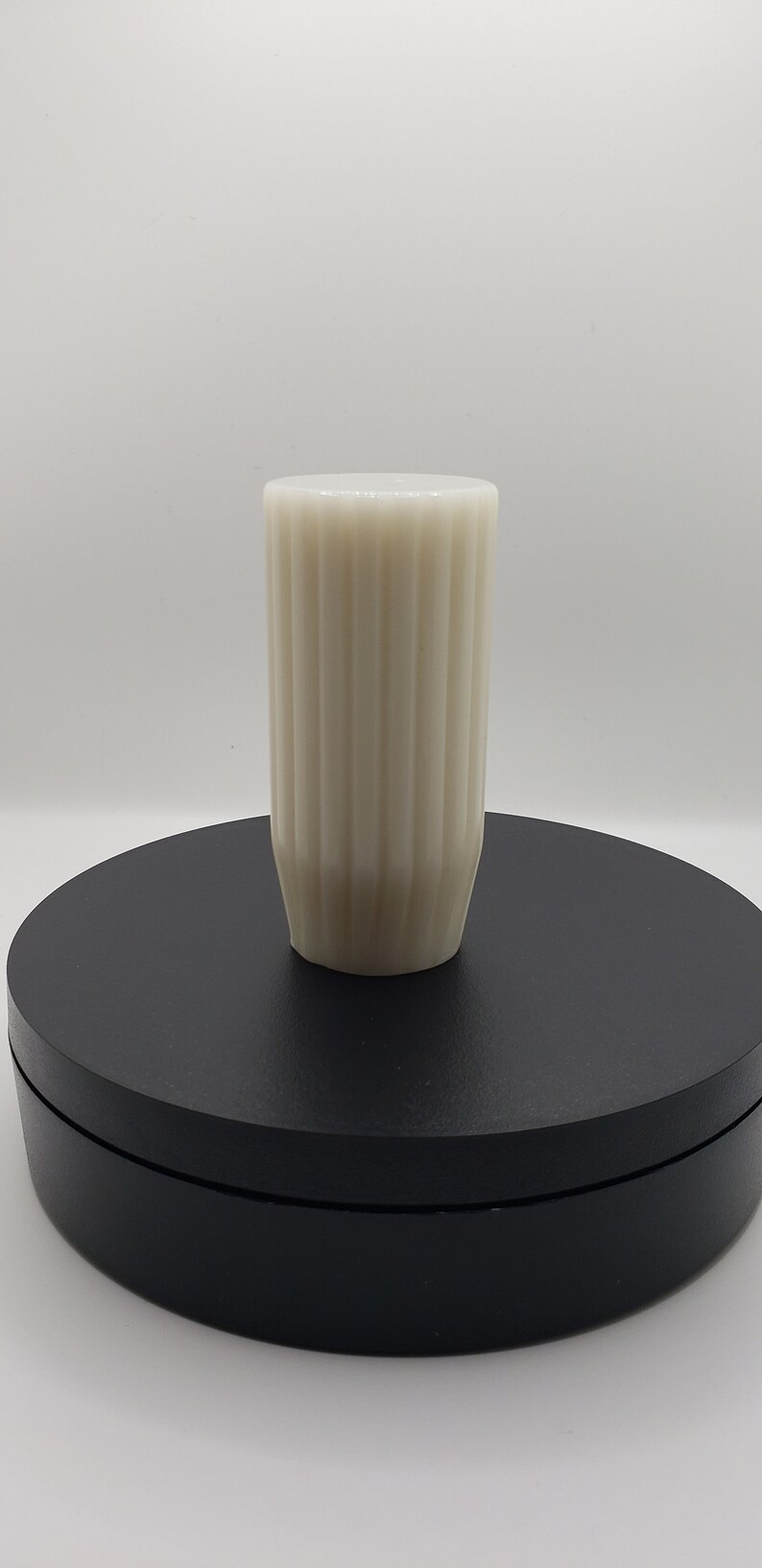 May include: A cylindrical off-white vase featuring a vertical ribbed texture along its exterior. The vessel has a flat top opening and a slightly tapered base, presenting a clean, uniform appearance with a consistent matte finish throughout.