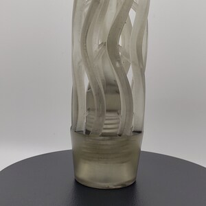 May include: A translucent, smoky gray cylindrical object featuring a cage-like structure composed of vertical, undulating, and spiraling ribbons. The base is solid and circular, transitioning into an open, airy design that tapers slightly toward the top.