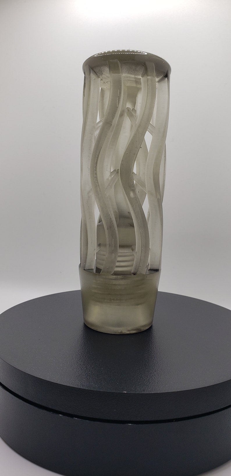 May include: A translucent, smoky gray cylindrical object featuring a cage-like structure composed of vertical, undulating, and spiraling ribbons. The base is solid and circular, transitioning into an open, airy design that tapers slightly toward the top.