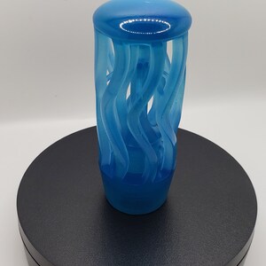 May include: A translucent blue bottle stopper featuring a cylindrical top with a smooth, rounded cap and a series of vertical, wavy, spiraling structural elements that connect the base to the top, creating an open, cage-like design.