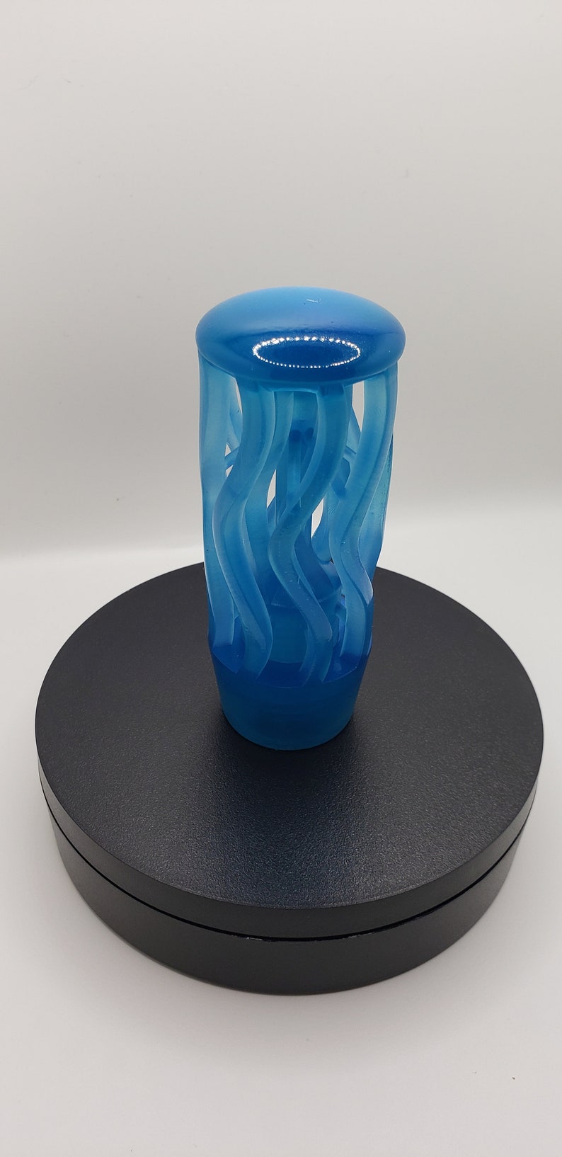 May include: A translucent blue bottle stopper featuring a cylindrical top with a smooth, rounded cap and a series of vertical, wavy, spiraling structural elements that connect the base to the top, creating an open, cage-like design.