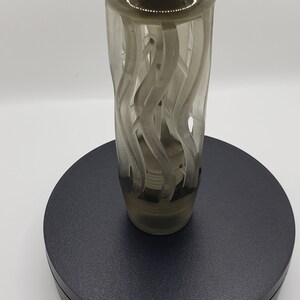 May include: A translucent, smoky gray gear shift knob featuring a cylindrical body with an intricate, open-work design of vertical, swirling, wave-like ridges. The top is smooth and rounded, while the base tapers to a solid, threaded attachment point.
