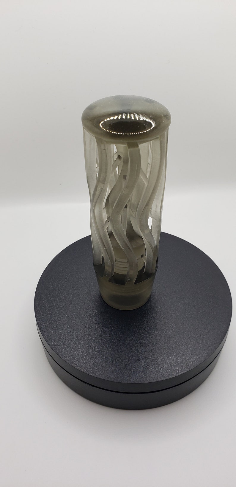 May include: A translucent, smoky gray gear shift knob featuring a cylindrical body with an intricate, open-work design of vertical, swirling, wave-like ridges. The top is smooth and rounded, while the base tapers to a solid, threaded attachment point.