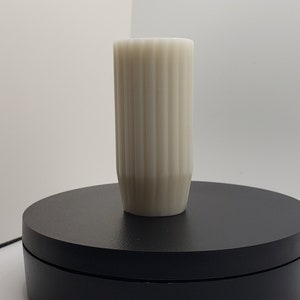 May include: A cylindrical off-white vase featuring a vertical ribbed texture along its exterior surface. The vessel has a straight, uniform body that tapers slightly toward the base, creating a clean and simple structural design for display.