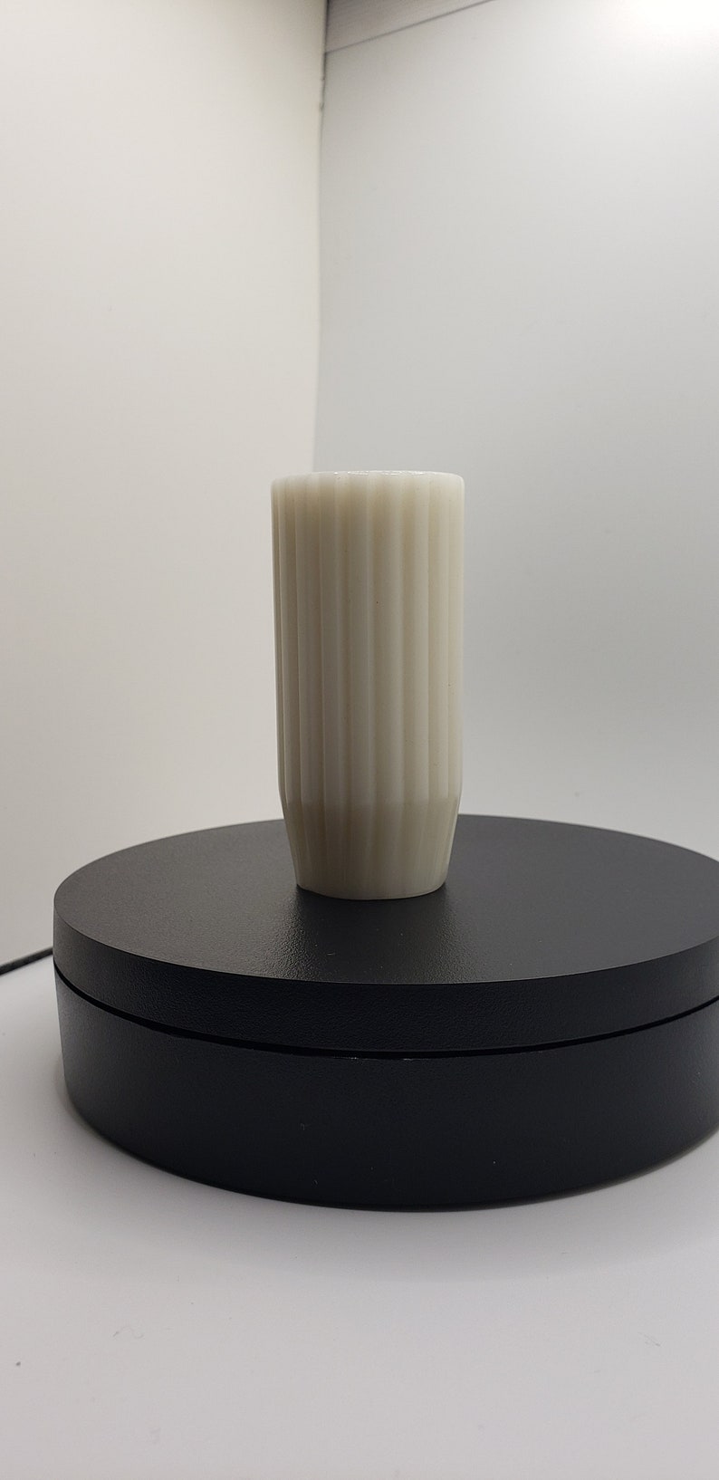 May include: A cylindrical off-white vase featuring a vertical ribbed texture along its exterior surface. The vessel has a straight, uniform body that tapers slightly toward the base, creating a clean and simple structural design for display.