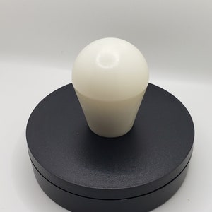 May include: A smooth, off-white, rounded knob with a tapered base sits centered atop a circular, matte black disc. The knob features a glossy finish that reflects light, while the black base has a flat top and a slightly recessed side profile.