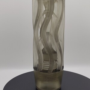 May include: A cylindrical, translucent gray gear shift knob featuring a series of vertical, wavy, raised ridges that wrap around the exterior. The base of the knob tapers slightly and includes a threaded interior section for attachment to a gear stick.