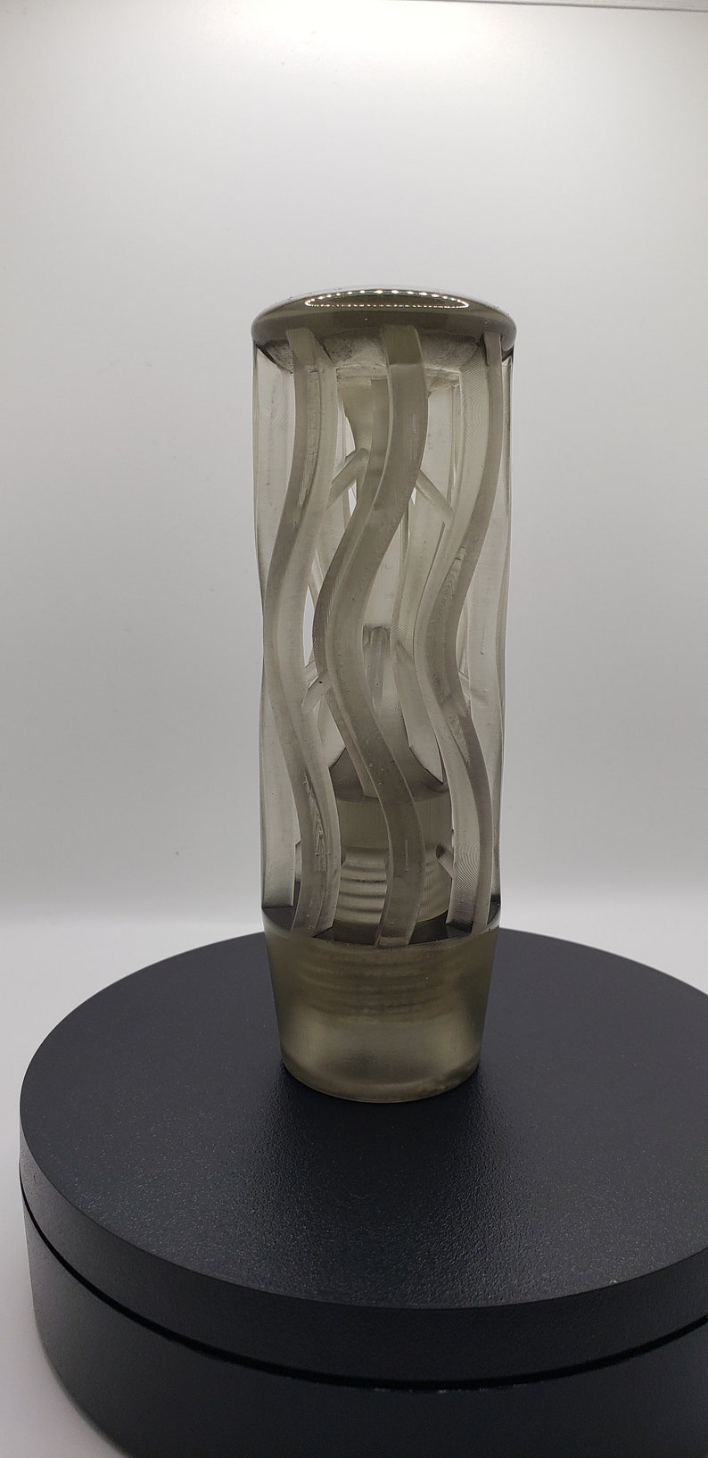 May include: A cylindrical, translucent gray gear shift knob featuring a series of vertical, wavy, raised ridges that wrap around the exterior. The base of the knob tapers slightly and includes a threaded interior section for attachment to a gear stick.