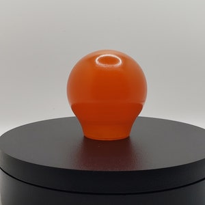 Könnte beinhalten: Ein glatter, kugelförmiger orangefarbener Knopf mit einer leicht verjüngten Basis. Die Oberfläche des orangefarbenen Objekts hat ein glänzendes Finish, das Licht reflektiert und einen hellen Glanzpunkt auf der oberen Kurve erzeugt.