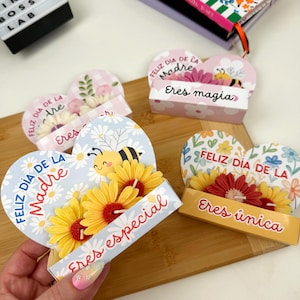 May include: Three heart-shaped paper cards with attached artificial flowers and bee illustrations. The cards feature the text Feliz Día de la Madre, Eres especial, Eres magia, and Eres única in colorful script and block lettering.