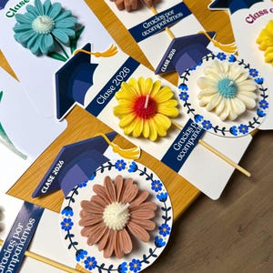 May include: A collection of flower-shaped candles attached to cardstock tags featuring a graduation cap illustration and the text "Clase 2026" and "Gracias por acompa&ntilde;arnos". The candles come in teal, yellow, and brown, each with a white center wick.
