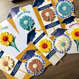 May include: A collection of graduation-themed party favors featuring flower-shaped candles attached to cardstock tags. The tags display the text "Clase 2026" and "Gracias por acompa&ntilde;arnos" alongside illustrations of graduation caps with yellow tassels.