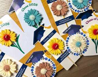 May include: A collection of graduation-themed party favours featuring flower-shaped candles attached to cardstock tags. The tags display the text "Clase 2026" and "Gracias por acompañarnos" alongside illustrations of graduation caps with yellow tassels.