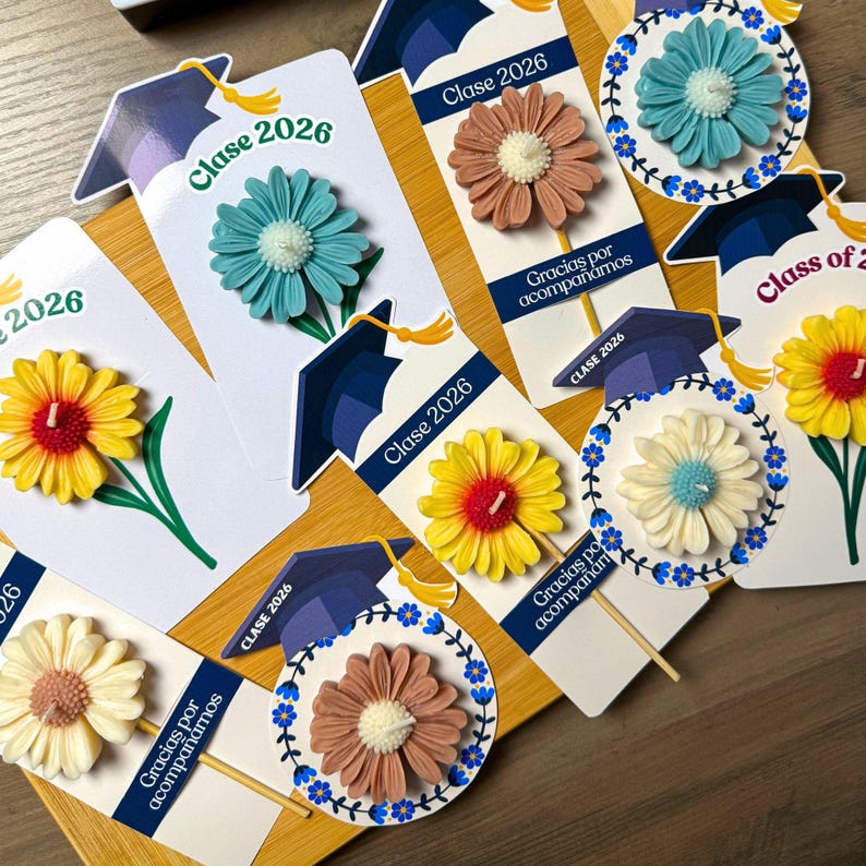 May include: A collection of graduation-themed party favors featuring flower-shaped candles attached to cardstock tags. The tags display the text "Clase 2026" and "Gracias por acompa&ntilde;arnos" alongside illustrations of graduation caps with yellow tassels.