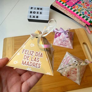 May include: A hand holding a yellow pyramid-shaped gift box with the text "FELIZ DÍA DE LAS MADRES" and small white flower designs. Two other gift boxes are visible, one with "FELIZ DÍA DE LA MADRE" and another with "Happy Mother's Day". A light box reads "PINK BOSS LAB".