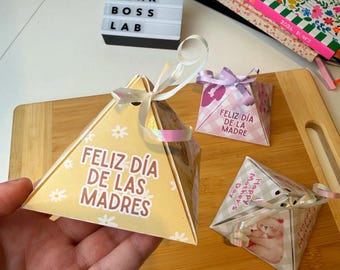 May include: A hand holding a yellow pyramid-shaped gift box with the text "FELIZ DÍA DE LAS MADRES" and small white flower designs. Two other gift boxes are visible, one with "FELIZ DÍA DE LA MADRE" and another with "Happy Mother's Day". A light box reads "PINK BOSS LAB".