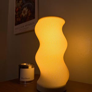 May include: A cylindrical table lamp with a wavy, ribbed exterior that emits a warm, golden glow. The lamp features a vertical, undulating pattern created by stacked layers, casting soft light from within its off-white, textured structure.