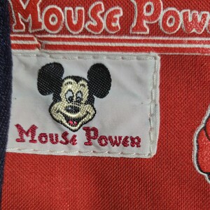 Może przedstawiać: Zbliżenie czerwonej tkaniny z napisem "Mouse Power" w kolorze białym i czerwonym. Biała naszywka przedstawia uśmiechniętą twarz Myszki Miki i napis "Mouse Power" w kolorze czerwonym.