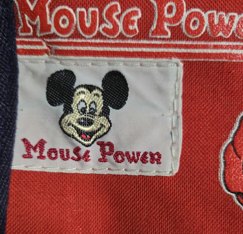 Może przedstawiać: Zbliżenie czerwonej tkaniny z napisem "Mouse Power" w kolorze białym i czerwonym. Biała naszywka przedstawia uśmiechniętą twarz Myszki Miki i napis "Mouse Power" w kolorze czerwonym.