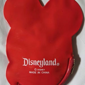 Może przedstawiać: Czerwona portmonetka w kształcie Myszki Miki z zapięciem na zamek błyskawiczny. Logo Disneyland jest wydrukowane na białym tle z przodu, wraz z prawami autorskimi Disneya i tekstem "Made in China".