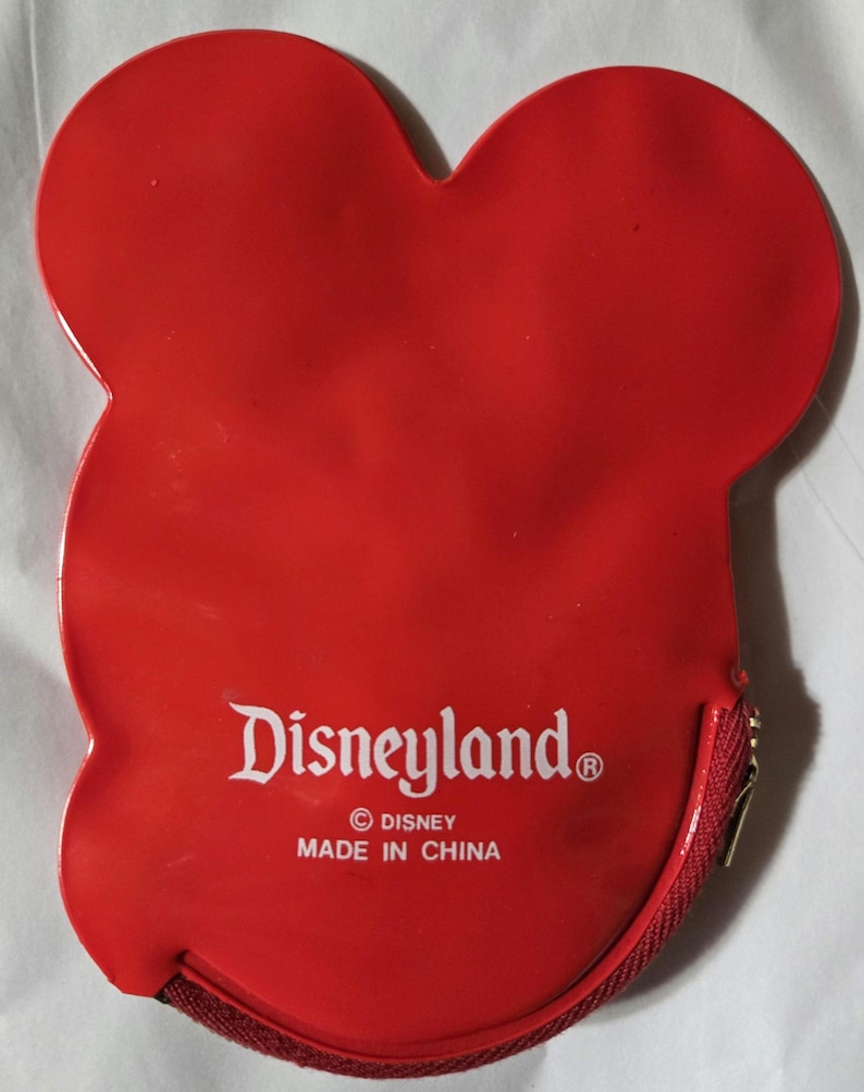 Może przedstawiać: Czerwona portmonetka w kształcie Myszki Miki z zapięciem na zamek błyskawiczny. Logo Disneyland jest wydrukowane na białym tle z przodu, wraz z prawami autorskimi Disneya i tekstem "Made in China".
