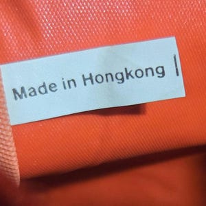 Może przedstawiać: Zbliżenie jasnoniebieskiej etykiety z czarnym napisem "Made in Hongkong". Etykieta umieszczona jest na tle żywego pomarańczowego tła o widocznej fakturze.