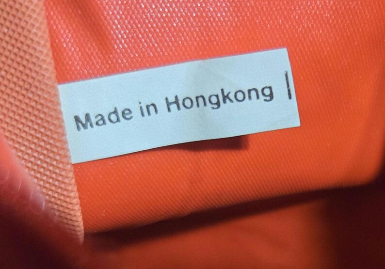 Może przedstawiać: Zbliżenie jasnoniebieskiej etykiety z czarnym napisem "Made in Hongkong". Etykieta umieszczona jest na tle żywego pomarańczowego tła o widocznej fakturze.