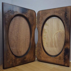May include: A hinged, wooden double photo frame with oval openings. The frame is stained a dark brown and features decorative, carved scrollwork in the corners. The frame is open, revealing the wooden backing and glass. The frame is designed to hold two photos.